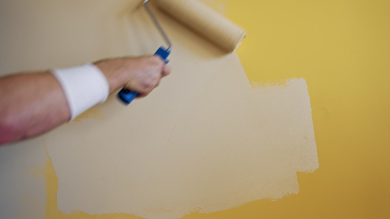 Roller painting yellow wall with creamy paint. Hand of a worker doing renovation the room with light color. Close-up. Makeover concept.