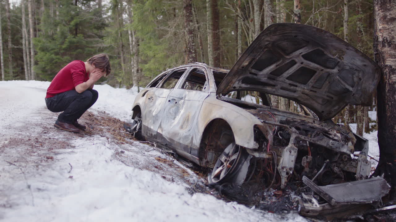 Crying young man crouching next to his written-off car wreck on side of icy road