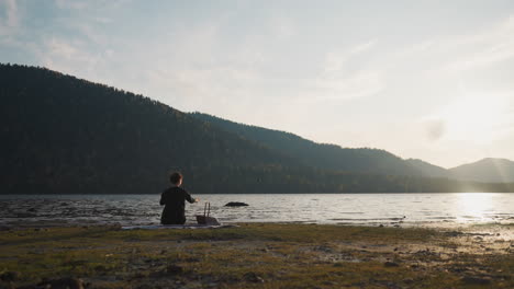 woman throws stones into lake on picnic. lady with food basket rests alone sitting on plaid after sunset. calm atmosphere on quiet riverbank in autumn