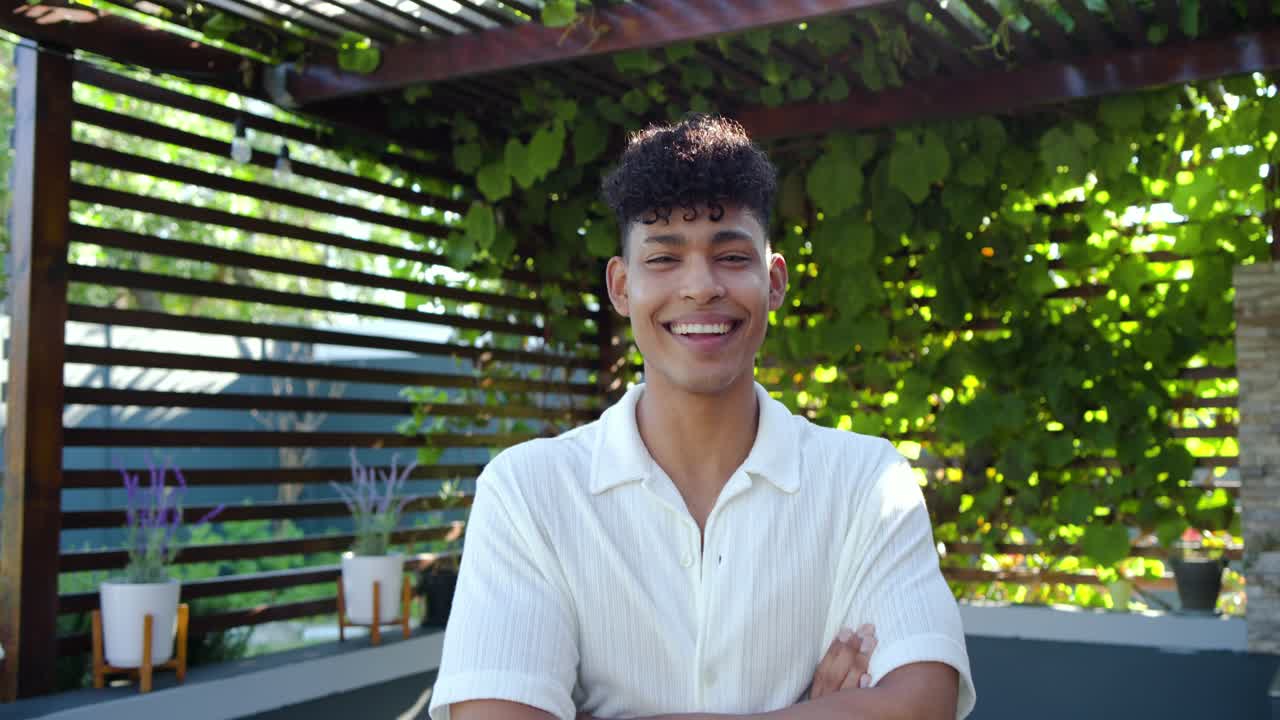 Afroameričan pod pergolou, se zkříženýma rukama, se narovnává a otáčí k fotoaparátu, usmívá se.