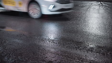 close-up view of a wet road at night, featuring light reflections on the glistening surface and two white cars driving by