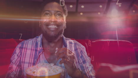 animation of letters over smiling african american man eating popcorn in cinema