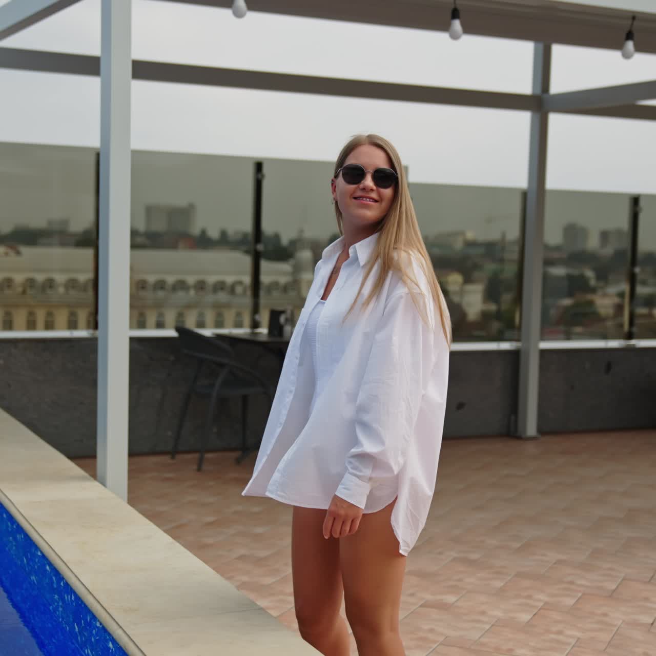 Young blonde lady in white swimsuit and shirt walks near the swimming-pool. Relaxing lounge zone at the roof of a building. Cityscape at backdrop