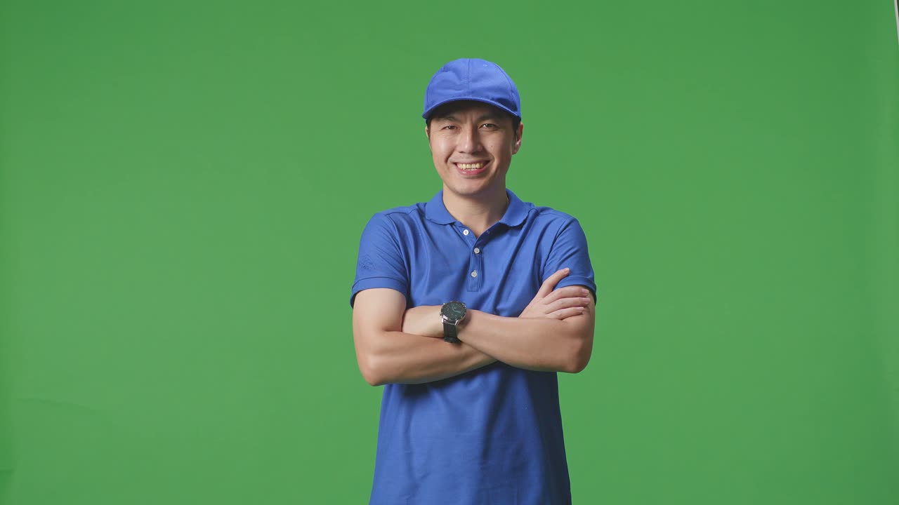 푸른 유니폼을 입은 아시아 남성 택배기사는 스튜디오의 녹색 스크린 배경에서 팔을 는 미소를 짓고 있습니다.