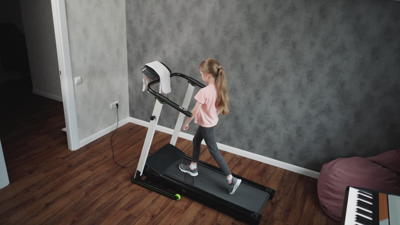 Working out session of young girl on treadmill holding handle for balance walking carefully in home fitness room with grey textured wall wooden floor background and keyboard visible behind