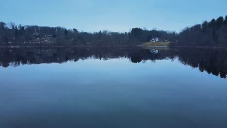 Drone-flying-just-above-the-water-of-a-calm-lake-with-a-glassy-surface-and-then-rising-in-the-air-to-show-a-small-town-and-mountains-after-sunset,-during-blue-hour-in-america