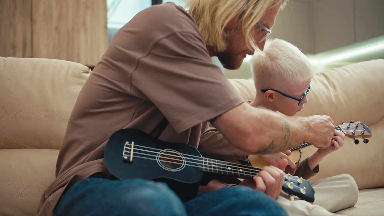 hombre rubio feliz con barba y gafas enseña a su pequeño hijo albino con gafas azules a tocar la guitarra correctamente y presionar acordes usando los dedos de su mano izquierda en el sofá en casa