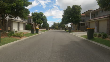 Street-view-of-apartment-houses-in-a-complex-with-trees-moving-in-the-wind-and-garbage-bins-outside-on-road