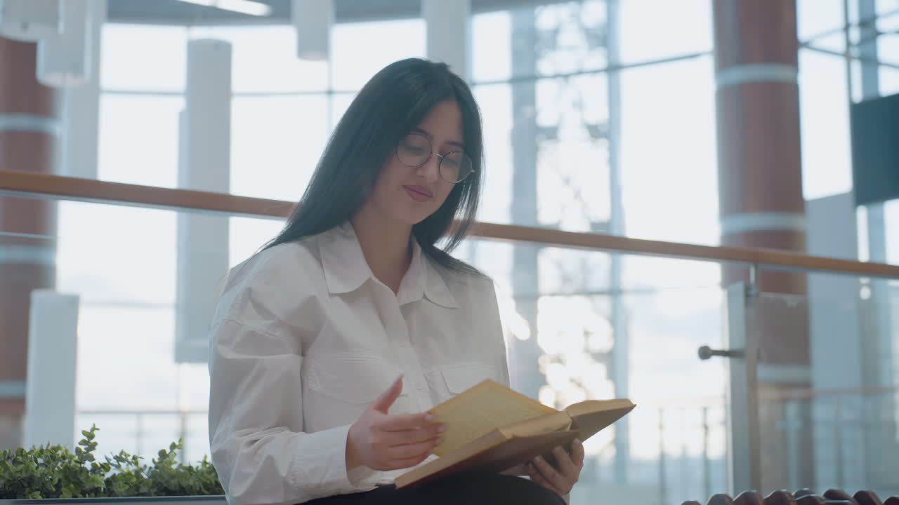 Girl seated indoors in bright modern mall, wearing glasses and white shirt, reads through open book with gentle warm smile, surrounded by greenery, soft lighting, and architectural glass background