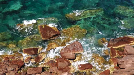 La-Cámara-Mira-Hacia-Abajo-En-Una-Costa-Rocosa-Con-Rocas-Rojas,-Agua-Cristalina-Y-Olas-Suaves