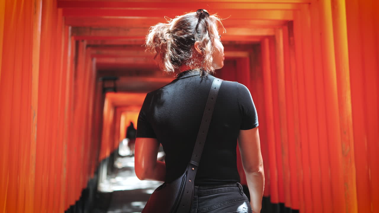mujer caminando a través de un túnel de puertas torii rojas, vista por detrás con el cabello atado y el traje negro