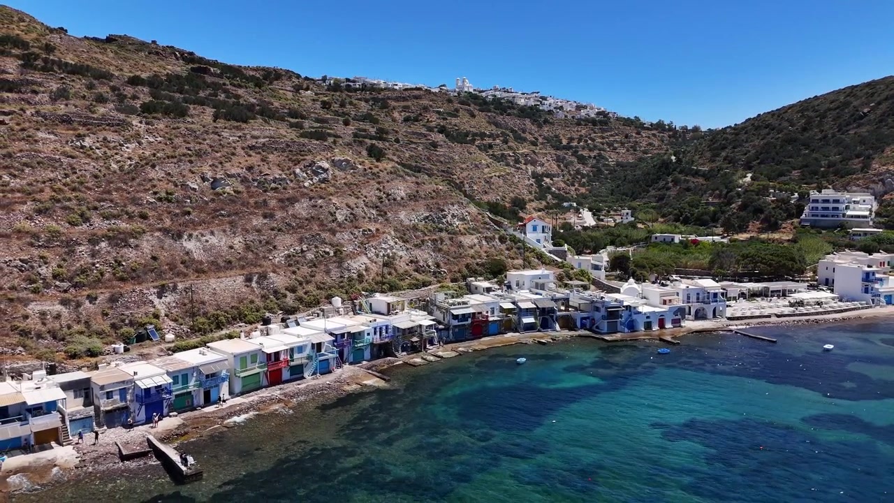 Il villaggio di pescatori di Klima presenta colorate case-barca syrmata lungo una costa rocciosa sotto colline aride, con piccole imbarcazioni sul Mar Egeo, Milos, Grecia, allontanamento con drone