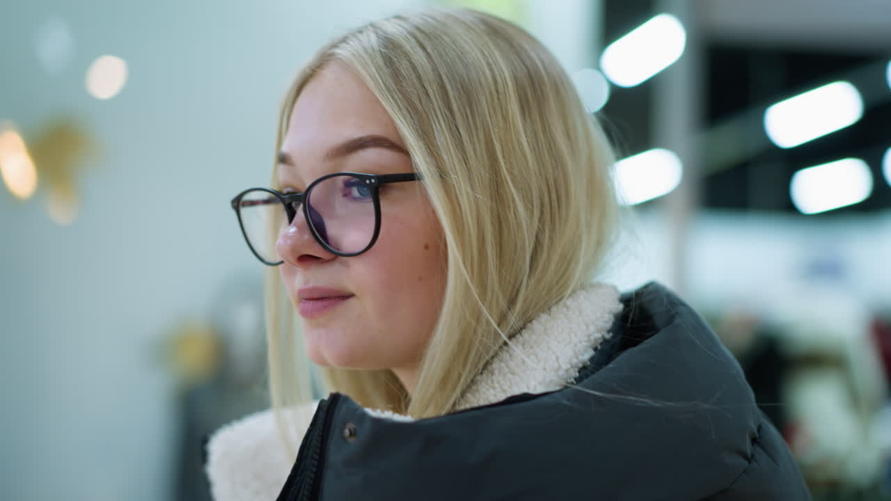 Close-up side view of student in black hoodie walking in well-lit mall, she looks calm as blurred background features retail space with people passing by, creating an atmosphere of relaxed shopping