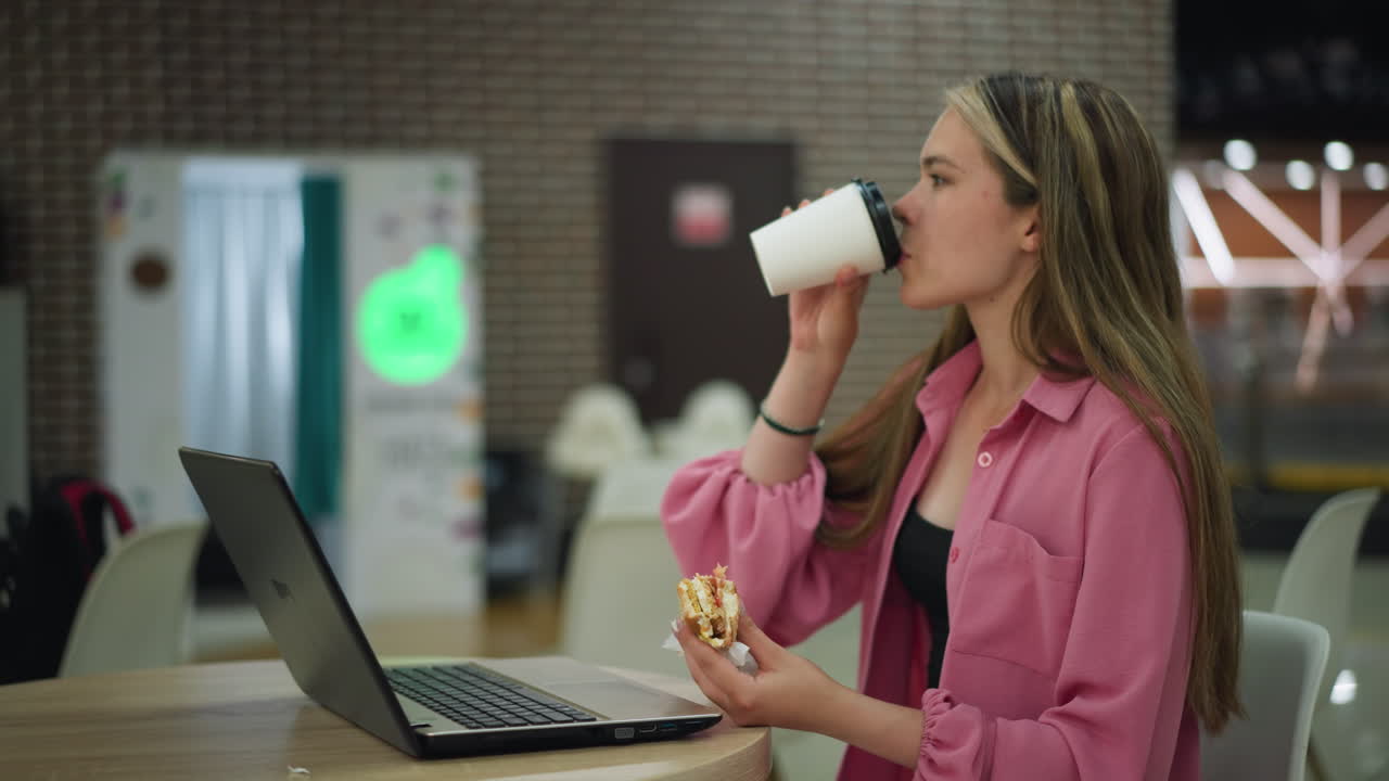 mujer sentada en un restaurante con una computadora portátil en frente, sosteniendo una hamburguesa en su mano izquierda mientras usa su mano derecha para tomar su bebida, el fondo está borroso con una luz de color suave parpadeando