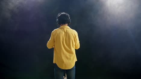 vista trasera de un niño con auriculares cantando en un micrófono condensador en el fondo negro de humo blanco