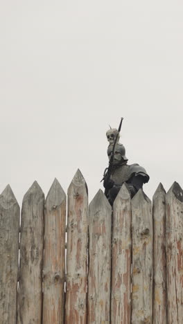 evil knight with spear walks near burnt wooden house behind fence rain. medieval demonic man looks for people in ancient village under grey sky low angle shot