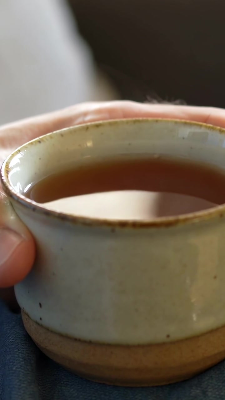 Video verticale: Una mano con la manica afferra il bordo di una tazza di ceramica sul divano, con vapore che sale per il calore.