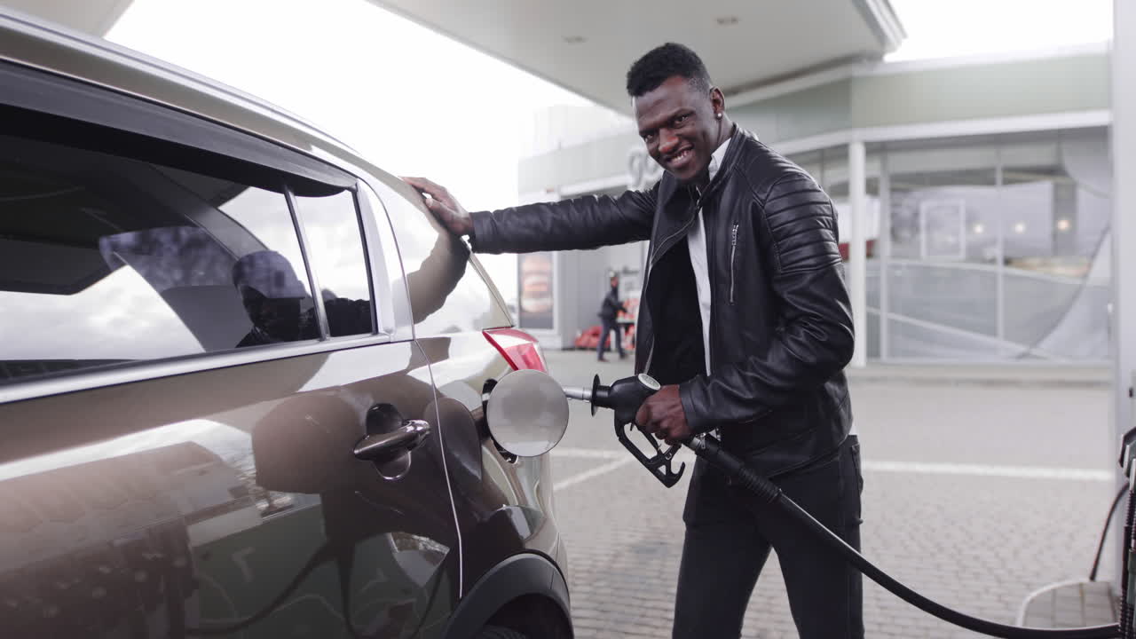 Giovane e allegro uomo d'affari afroamericano che fa rifornimento alla sua moderna auto di lusso in una stazione di servizio, sorridendo