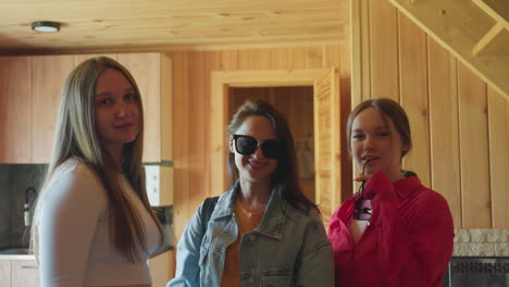 three elegant women stand close together inside cozy wooden interior as lady in red shirt lifts sunglasses while others pose calmly with confident expressions in soft natural lighting