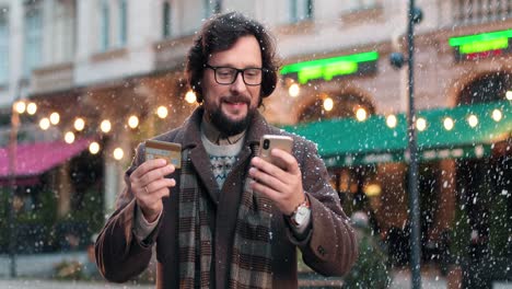 close-up view of caucasian man wearing eyeglasses shopping online using smartphone on the street while it‚äôs snowing in christmas