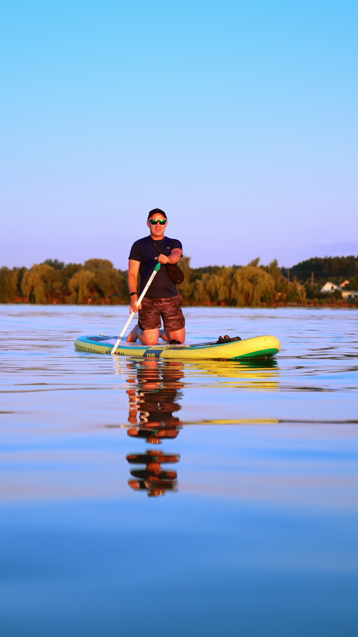 Practicing water sport on the river. Man paddle boarding solo sitting on the sup board. Vertical video.