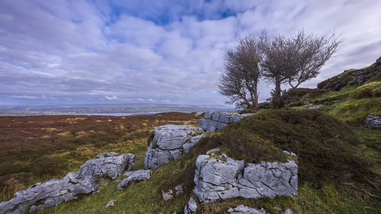 Panorama-timelapse af et landligt landskab med træer og klipper i forgrunden, græsbevokset moseområde og bakker med sø i det fjerne på en skyet dag, set fra Carrowkeel i County Sligo i Irland