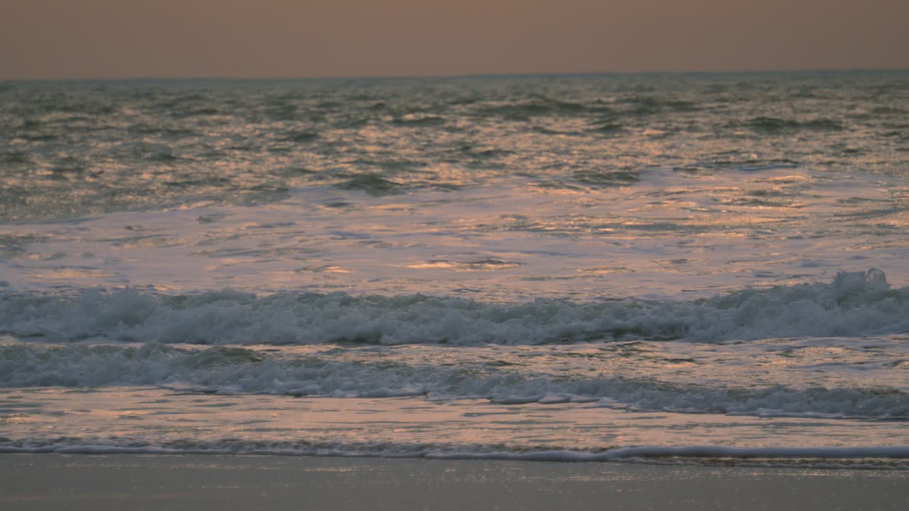 Slow Motion Ocean Footage with Orange Sunrise Lighting Reflecting on Surface as Small Waves Roll onto Beach. ProRes 4K.
