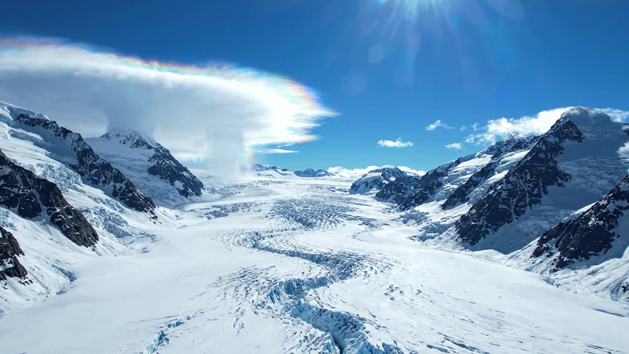 En fantastisk flyfoto av majestetiske snødekte fjell og en vidstrakt isbre under en lys blå himmel med tynne skyer og strålende sollys.