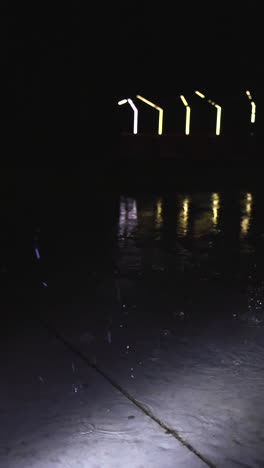 vertically filmed close up video of raindrops falling on a gray concrete wet and water covered slab or ground in slow motion. water is splashing around during night time and street lights in the back