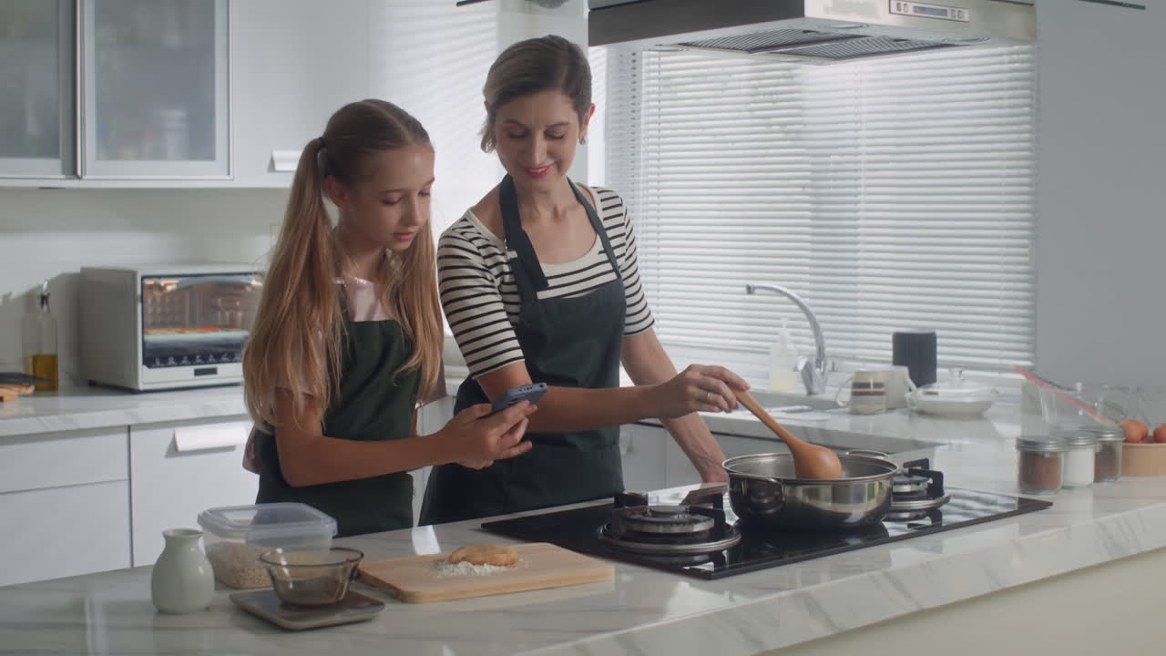 Teen Girl Showing Something on Smartphone to Mother Cooking at Kitchen