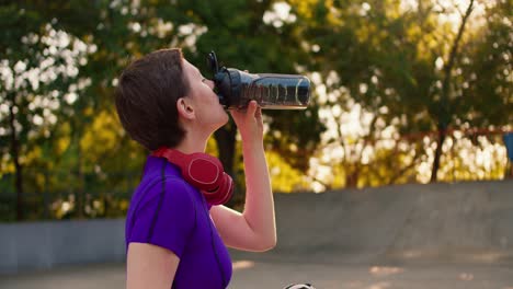 side view of a girl in a purple top and red headphones in a short haircut drinks special water in a skate park in summer