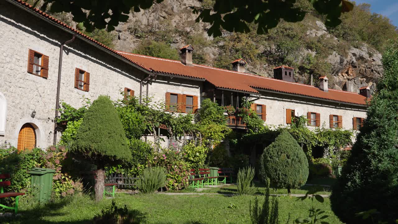 A peaceful shot of the historic Morača Monastery in Montenegro, nestled by a rocky cliff. The shot shows the traditional stone residential buildings and lush inner garden