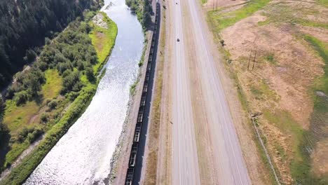 Toma-Aérea-De-Un-Tren-Que-Circula-Por-Una-Carretera-Y-Un-Río-En-La-Costa-Oeste-De-EE.UU.