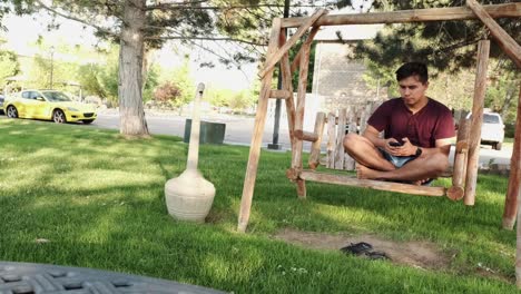 young handsome caucasian male relaxing on wooden bench using cellphone as cute grey cats approach him in public park