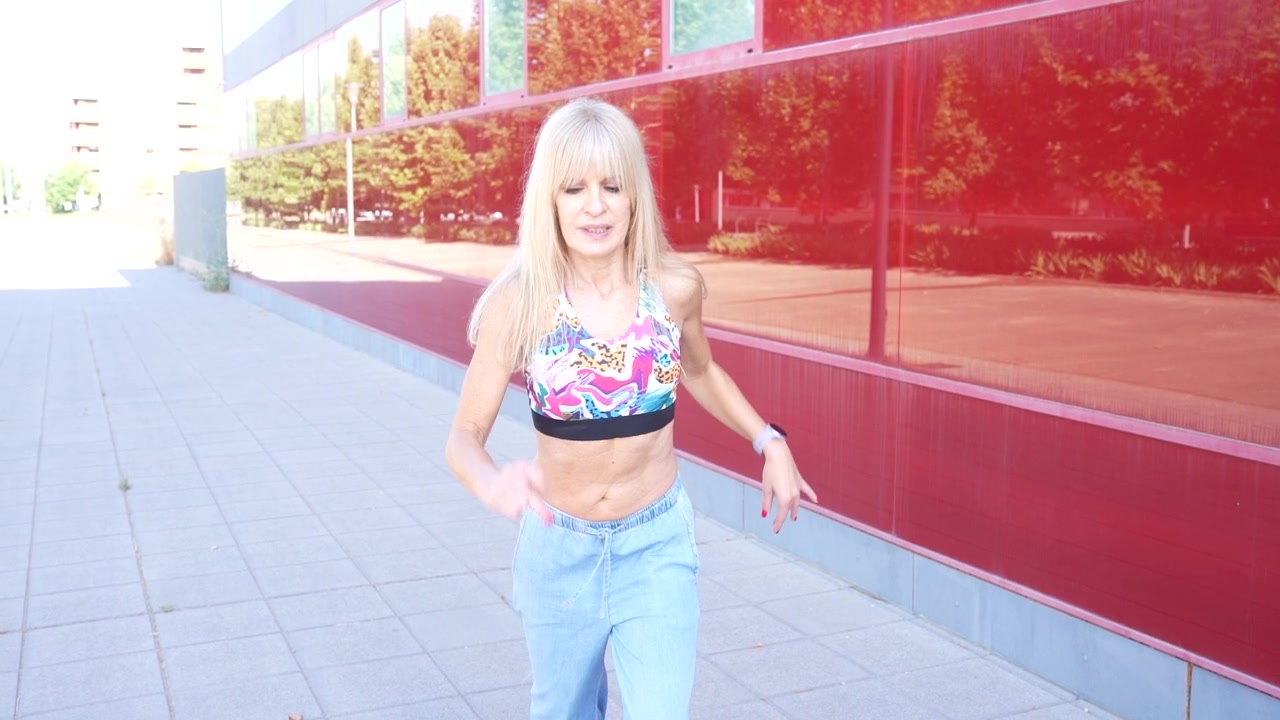 En blond kvinde iført en farverig top danser udendørs med en moderne rød væg som baggrund.