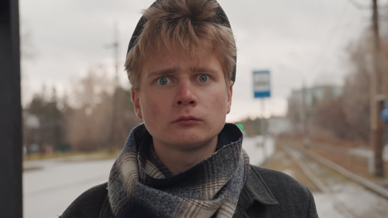 Young white creator at bus stop, overcast morning with wool cap and patterned scarf, sideways glances toward passing traffic, cold commuter atmosphere, cinematic portrait emphasizing solitude