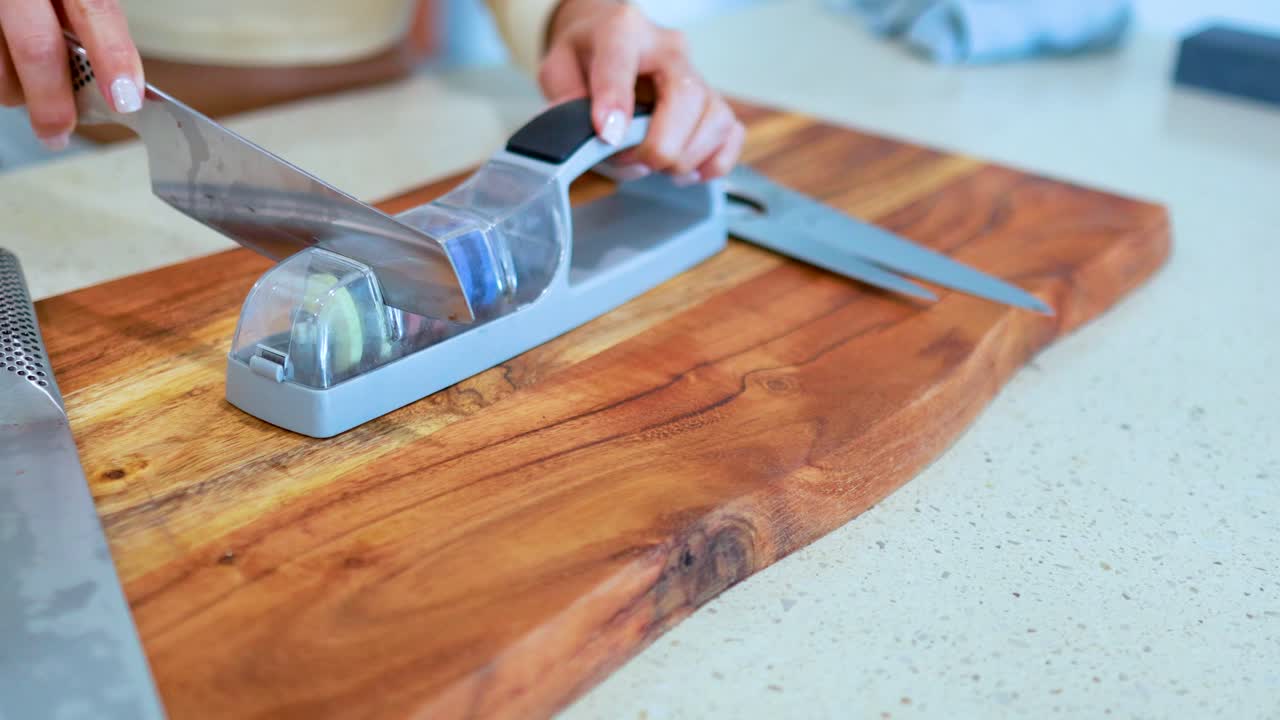Person sharpens a chef’s knife using a handheld whetstone sharpener on a wooden cutting board in a well-lit kitchen, captured in close-up