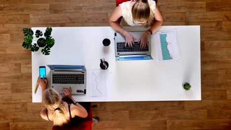 fotografía superior, estado de ánimo de trabajo relajado, dos chicas, escribiendo en teléfonos inteligentes, comprobando el teléfono con pantalla azul, mientras están sentados en un escritorio común blanco interior