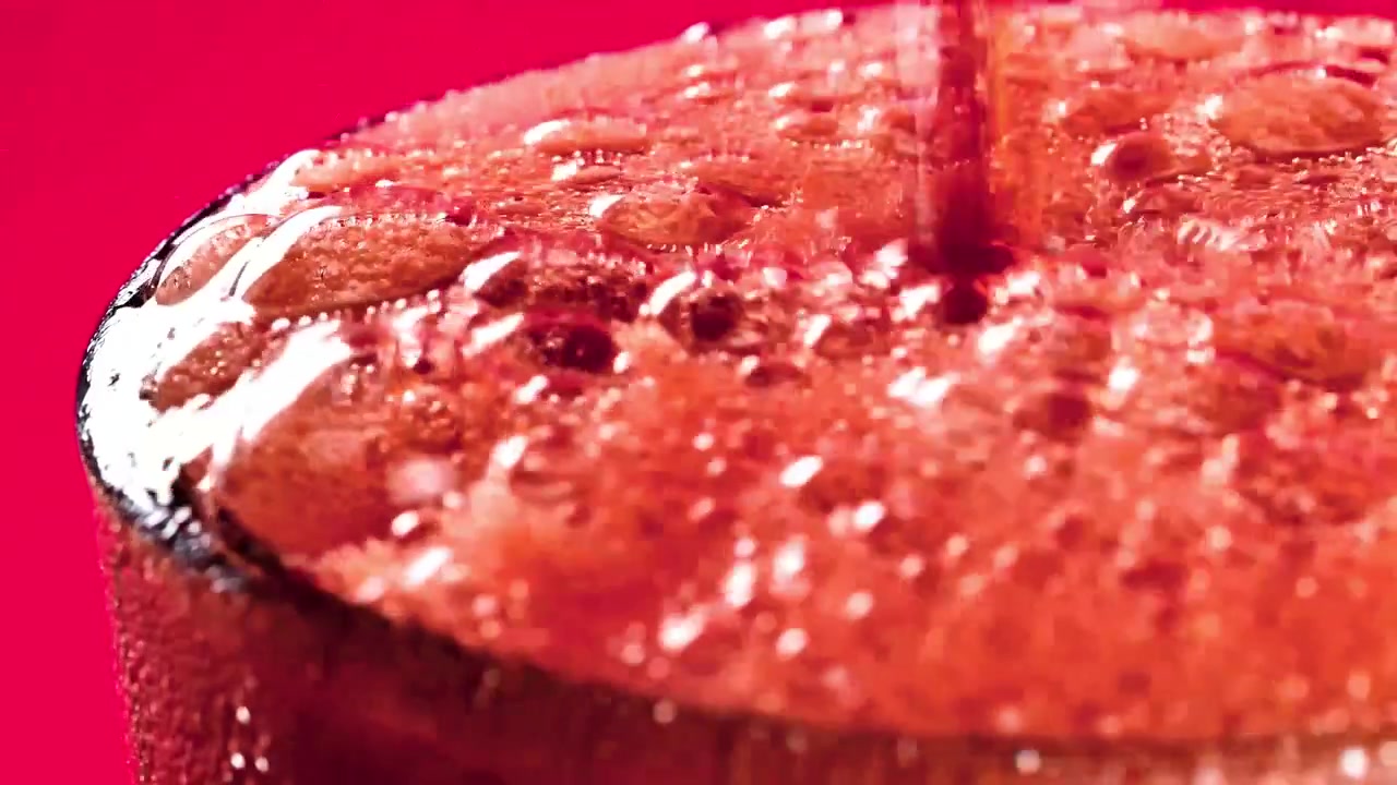 Makronærbillede af kold cola, der hældes over isterninger i et klart glas, hvilket skaber brusende bobler, skum og små stænk mod en levende rød studiebaggrund.
