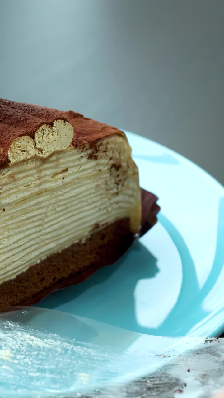 Mani rimuovono con cura la pellicola trasparente da una fetta di torta tiramisù a strati su un piatto blu, con illuminazione naturale.