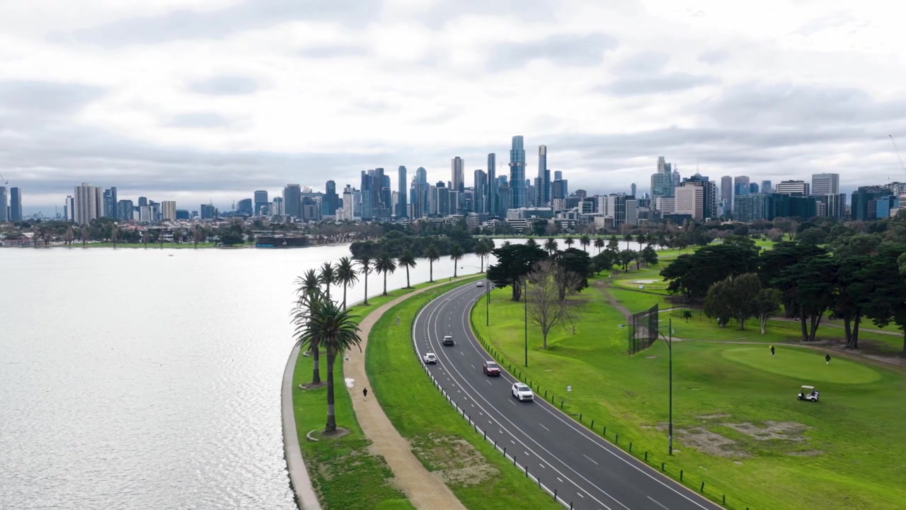Krásné filmové letecké záběry z dronu jezera Albert Park, ukazující klikatou silnici podél jezera s projíždějící dopravou, cyklisty, palmami, rozlehlými parky a panoramatem centra Melbourne v pozadí.