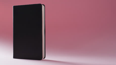 close up of closed black book standing vertical with copy space on pink background in slow motion