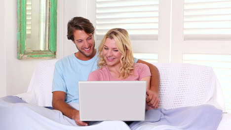 pareja sentada en el sofá bajo una manta usando una computadora portátil