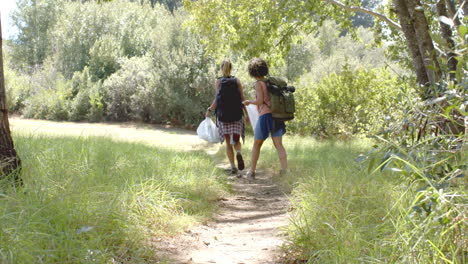 two women walk along a narrow trail in a lush, green forest setting, collecting trash