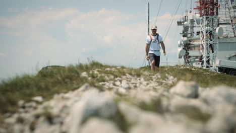 hiker with a cap and an orange backpack walking away from the radio tower on rocky path with patches of grass