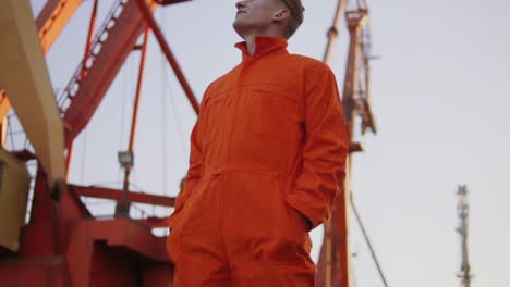 Apuesto-Joven-Trabajador-De-Almacén-De-Contenedores-Con-Uniforme-Naranja-Parado-Junto-Al-Barco-En-El-Puerto-Y-Mirando-Hacia-Arriba.-Gran-Grúa-En-El