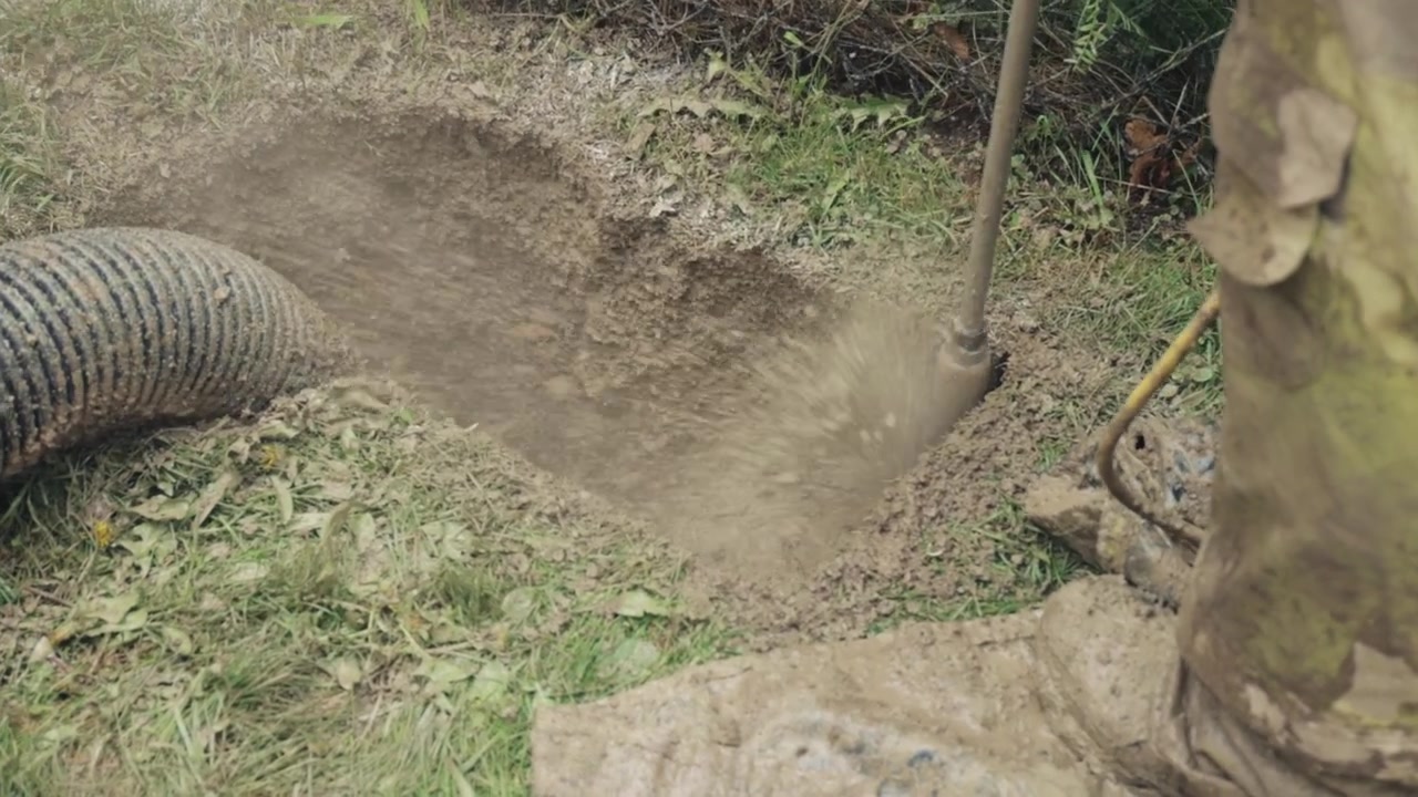 Lo scavo idrovacuum espone un foro per palo su un ciglio erboso mentre le squadre preparano il sito per la fondazione del palo dell'illuminazione stradale e i lavori di canalizzazione; sono visibili il tubo di aspirazione e la fanghiglia di terra, sottolineando l'uso sicuro.