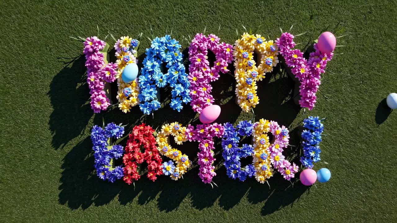 Aerial view of 'Happy Easter!' spelled with colorful flowers on grass, surrounded by eggs