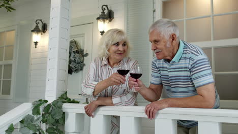 Senior-elderly-caucasian-couple-drinking-wine,-looking-ahead,-making-a-kiss-in-porch-at-home