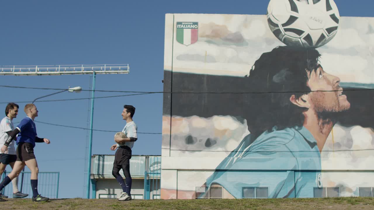 A young soccer player admires a mural of maradona, catching a ball and joining friendly teammates